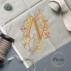 May include: A gold embroidered letter N with ornate flourishes and a beaded design on a gray linen fabric background. The fabric has a lace trim and the word "Pteris" is written in a gold circle on the bottom right corner.