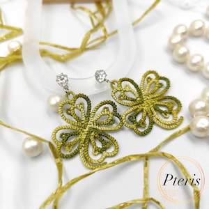 May include: A pair of handmade, green and yellow clover-shaped earrings. Each earring features a sparkling, clear gem at the top and a dangling, openwork design. The earrings are displayed with pearls and gold ribbon.
