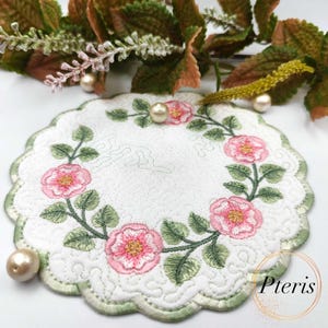 May include: A white, scalloped-edge coaster with an embroidered floral design. Pink roses and green leaves form a circular pattern. The coaster has a quilted texture and a green border. Faux pearls and greenery are in the background.