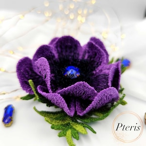 Può includere: Un fiore viola di peluche fatto a mano, con un centro scuro e un dettaglio blu a forma di gemma. Il fiore ha foglie e steli verdi. L'immagine è su uno sfondo bianco con luci soffuse e sfocate.