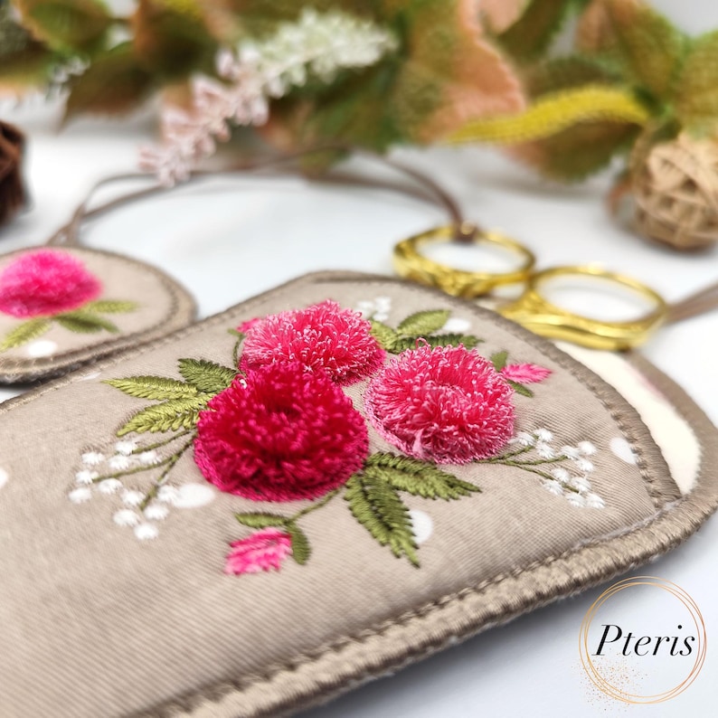 K&ouml;nnte beinhalten: Eine beige Stoffschere-H&uuml;lle mit rosa gestickten Blumen und wei&szlig;en Punkten. Die H&uuml;lle hat eine braune Schnur und einen goldenen Scherenhalter aus Metall.