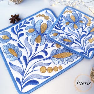 Puede incluir: Dos posavasos de tela bordados con un diseño floral azul y dorado. Los posavasos tienen un fondo blanco y un borde azul.