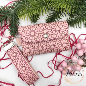 以下が含まれることがあります： 明るいピンク色の財布とリストレットキーフォブのセット。どちらのアイテムも、濃いピンク色の糸で刺繍された幾何学的な星模様が特徴です。財布はスナップ留めです。キーフォブには、シルバーカラーの金属製クラスプが付いています。