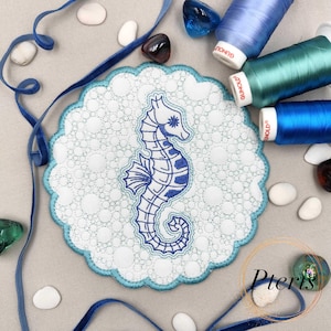 Op de afbeelding: Een ronde witte stoffen patch met een geschulpte lichtblauwe rand en een geborduurd blauw zeepaardje. De patch is omringd door blauw lint, drie klossen blauw en groen garen en verschillende gladde witte steentjes.