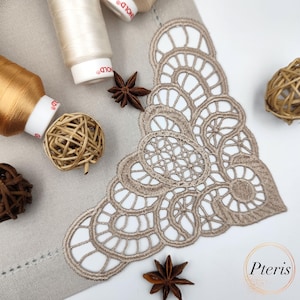 May include: A beige lace corner design embroidered on a light brown linen fabric. The design features intricate circles and swirls. There are spools of thread and star anise pods on the fabric.