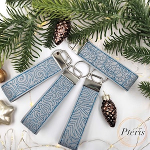 May include: A collection of keychains with embroidered designs in blue thread on a gray background. Each keychain has a silver metal clasp. The keychains are arranged with Christmas decorations, including pine needles and ornaments.