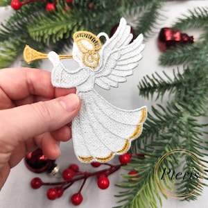 May include: Embroidered white angel ornament with gold accents, holding a trumpet. The angel has detailed wings and a flowing gown. The ornament is likely for Christmas or holiday decor.