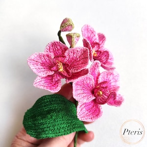 Puede incluir: Una flor de orqu&iacute;dea bordada de color rosa con una hoja verde. La flor est&aacute; hecha de tela y tiene un aspecto delicado y realista.