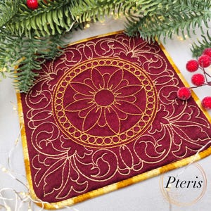 May include: A square, burgundy coaster with a gold floral design. The coaster features a central flower motif surrounded by a circular pattern and ornate, swirling details. The edges are trimmed with gold fabric. The word "Pteris" is in the bottom right corner.