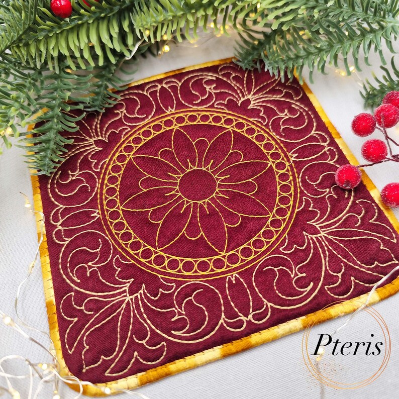 May include: A square, burgundy coaster with a gold floral design. The coaster features a central flower motif surrounded by a circular pattern and ornate, swirling details. The edges are trimmed with gold fabric. The word "Pteris" is in the bottom right corner.