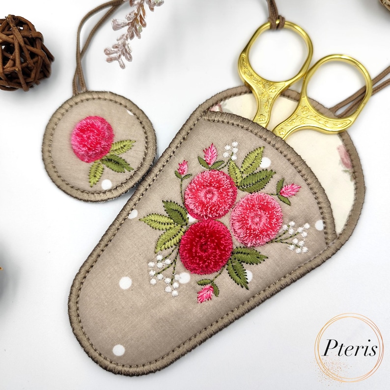 K&ouml;nnte beinhalten: Ein beigefarbener Stoffschneider mit rosa gestickten Blumen und wei&szlig;en Punkten. Der Schneider hat eine Schlaufe zum Aufh&auml;ngen und eine goldene Schere im Inneren.