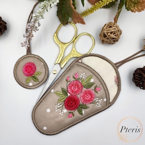 K&ouml;nnte beinhalten: Ein brauner Stoffschneider mit einem floralen Stickmuster mit rosa Blumen und gr&uuml;nen Bl&auml;ttern. Der Schneider hat ein wei&szlig;es Punkte-Muster und einen kleinen runden Stoffkreis mit einem rosa Blumenstickmuster. Eine goldene Schere ist in den Schneider gelegt.