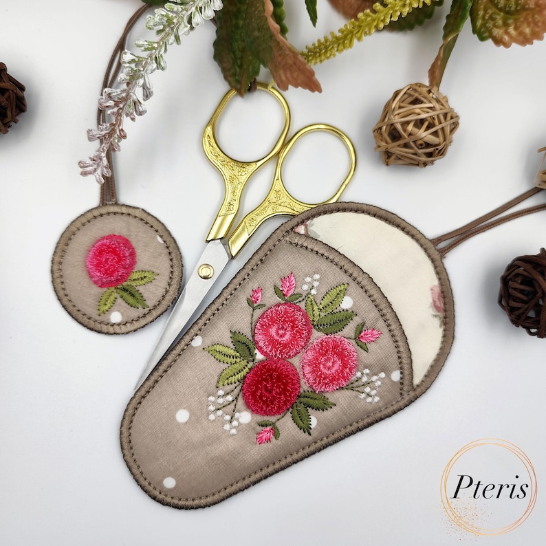 K&ouml;nnte beinhalten: Ein brauner Stoffschneider mit einem floralen Stickmuster mit rosa Blumen und gr&uuml;nen Bl&auml;ttern. Der Schneider hat ein wei&szlig;es Punkte-Muster und einen kleinen runden Stoffkreis mit einem rosa Blumenstickmuster. Eine goldene Schere ist in den Schneider gelegt.