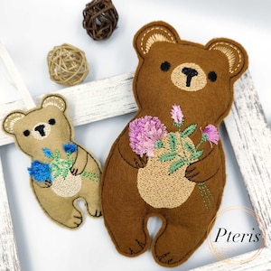 May include: Two felt bear ornaments. One is light brown with blue embroidered flowers, the other is dark brown with purple embroidered flowers. Both have stitched details and black eyes. The ornaments are on a white background.