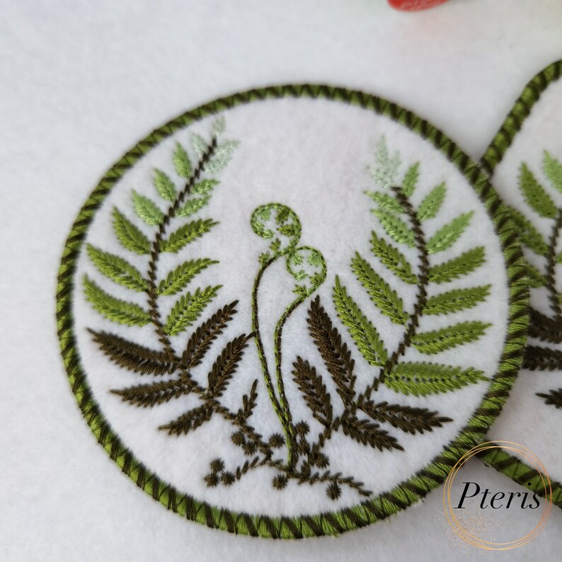 May include: A white felt circle with a green embroidered fern design. The fern has dark green stems and light green leaves. The circle has a green embroidered border.