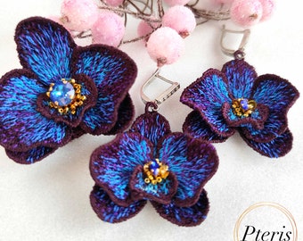 May include: A pair of embroidered orchid earrings and a matching brooch in deep purple and vibrant blue thread. Each piece features a central blue crystal surrounded by gold beads. The set is displayed with pink decorative berries and the text Pteris.