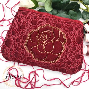 May include: A burgundy zipped pouch featuring a gold embroidered rose. The pouch has a textured pattern of small circles and the word "Pteris" in a cursive font.