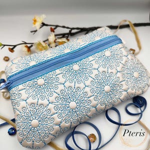 May include: A white zippered pouch with a blue zipper and a blue and white swirling pattern. The pouch is lying on a table with a blue ribbon and a branch with white flowers.
