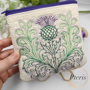 May include: Cream-coloured zippered pouch featuring an embroidered thistle design in green, purple, and black. The pouch has a quilted texture and a purple zip. The brand name "Pteris" is visible.