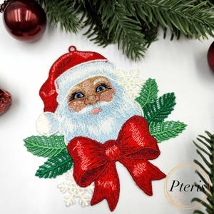 Può includere: Ornamento natalizio ricamato di Babbo Natale con cappello e fiocco rossi, occhi blu e barba bianca. L'ornamento è circondato da foglie verdi e fiocchi di neve. La parola "Pteris" è visibile in basso a destra.