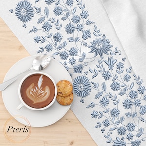 Puede incluir: Un camino de mesa de lino blanco con un diseño floral bordado en azul. Un platillo blanco con una taza de café y dos galletas está sobre una mesa de madera.