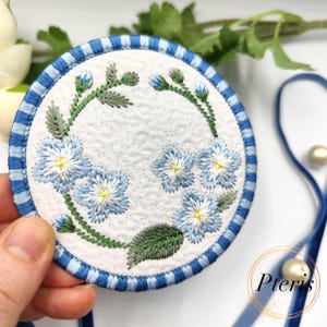 May include: A round, white coaster with a blue and white striped border. The coaster features embroidered blue flowers with yellow centers and green leaves. The design is centered on a quilted background.