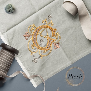 May include: A gold embroidered letter G with ornate floral details on a gray linen fabric. The letter is surrounded by a decorative border of small gold beads.
