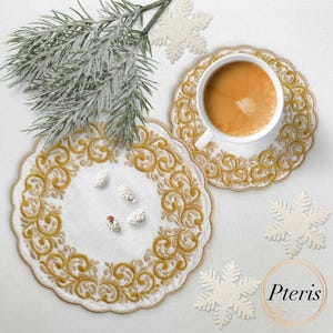 Puede incluir: Dos posavasos blancos bordados con un diseño floral dorado. Los posavasos están sobre una superficie blanca con una rama de pino y copos de nieve.