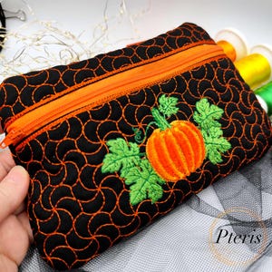 May include: Black zippered pouch with an embroidered orange pumpkin and green leaves. The pouch has an orange zipper and stitching. The background is black with an orange swirl pattern. The pouch is a fall-themed accessory.