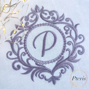 Puede incluir: Un monograma bordado en morado con la letra "P" en un marco decorativo. El marco está hecho de diseños florales en espiral.