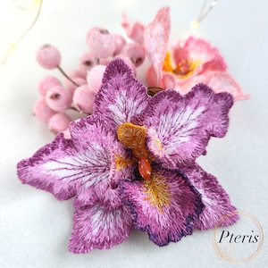 Puede incluir: Primer plano de una flor de orquídea bordada en morado y blanco con una flor bordada en rosa y blanco en el fondo. Las flores están rodeadas de pequeñas cuentas rosas y blancas.
