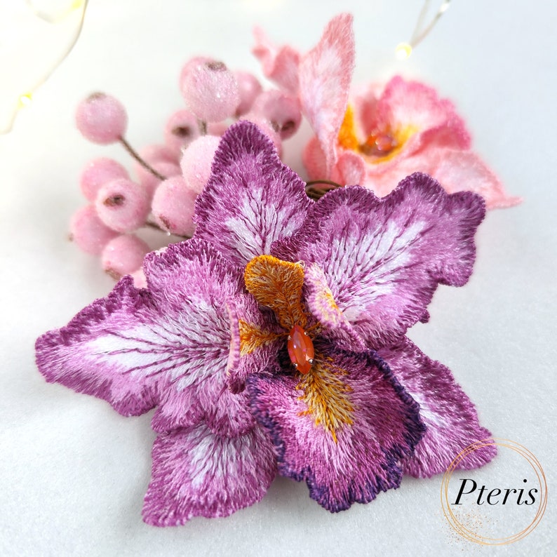 Puede incluir: Primer plano de una flor de orqu&iacute;dea bordada en morado y blanco con una flor bordada en rosa y blanco en el fondo. Las flores est&aacute;n rodeadas de peque&ntilde;as cuentas rosas y blancas.