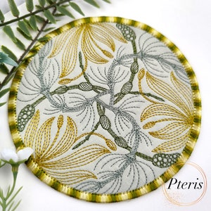 Puede incluir: Un posavasos redondo bordado con un diseño floral en tonos amarillos y verdes. El posavasos tiene un borde verde y amarillo.