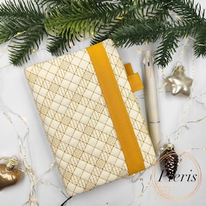 May include: Cream-colored journal with a geometric gold pattern and a mustard-yellow elastic band. A matching pen and star-shaped ornament are next to the journal. The background includes Christmas tree branches and decorative lights.