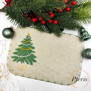 Puede incluir: Un mantel individual rectangular de color blanquecino con un borde verde presenta un árbol de Navidad bordado adornado con luces de cadena amarillas y blancas. El mantel individual está decorado con un patrón sutil y repetitivo. Adornos navideños y vegetación rodean el mantel individual.