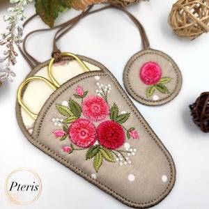 K&ouml;nnte beinhalten: Ein beigefarbener Stoffschneider mit einem floralen Stickmuster mit rosa Rosen und gr&uuml;nen Bl&auml;ttern. Der Schneider hat ein wei&szlig;es Punkte-Muster und eine braune Schnur zum Aufh&auml;ngen. Der Schneider ist mit einer goldenen Schere gef&uuml;llt.