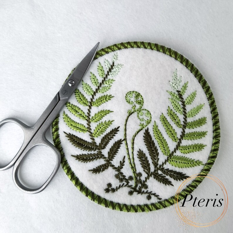 May include: A round, white felt coaster with green embroidery of fern leaves and a fern frond with a cluster of round, green spores. The coaster has a green stitched border. The word "Pteris" is written in green below the coaster.