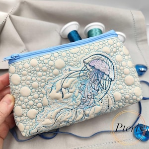 May include: A light blue zippered pouch with a detailed embroidered jellyfish design. The pouch features a textured, dotted pattern and a light blue zipper. The jellyfish is rendered in shades of blue and purple, with delicate tendrils.
