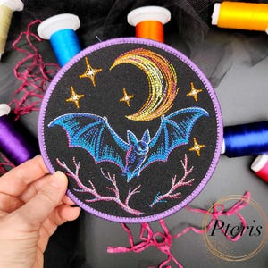 Può includere: Una toppa circolare in tessuto nero con un ricamo raffigurante un pipistrello blu in volo sotto una mezzaluna gialla e arancione e quattro stelle dorate, con rami rosa sul fondo e un bordo cucito di colore viola.