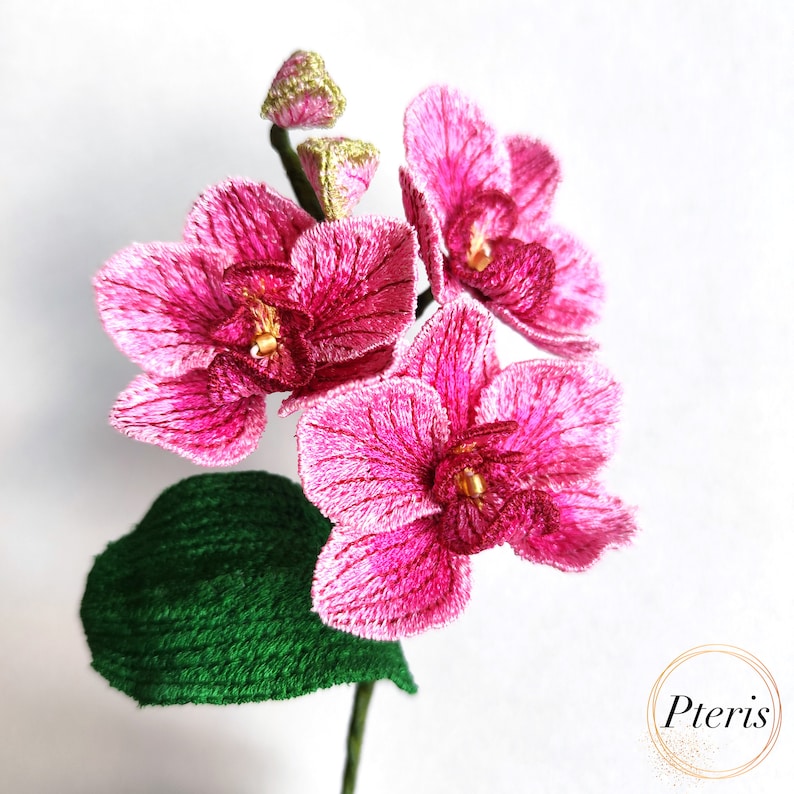 Puede incluir: Una flor de orqu&iacute;dea rosa con una hoja verde, bordada con detalles finos. La flor es una opci&oacute;n popular para la decoraci&oacute;n del hogar y los regalos.
