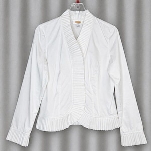 May include: A white long-sleeve jacket with a pleated design on the front and cuffs. The jacket has a V-neck and is hanging on a clear hanger. The jacket is made of a lightweight fabric.