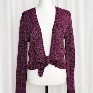 May include: A deep magenta and black knit cardigan featuring a cable knit pattern and a textured stitch design. The garment has long sleeves, a V-neckline, and a front tie closure at the waist, displaying a two-tone color effect throughout the fabric.