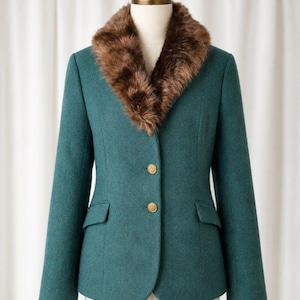 Pode incluir: Um blazer verde-azulado com gola de pelo sintético castanho, mangas compridas e dois botões dourados na frente. O casaco inclui dois bolsos com aba na parte inferior e é exibido num manequim contra um fundo de cortina branca.