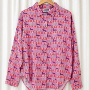 May include: A long-sleeve button-down shirt in a pink base color featuring a repeating pattern of purple horses and vertical red and white chevron stripes. The shirt has a standard collar, a front button placket, and a curved hemline.