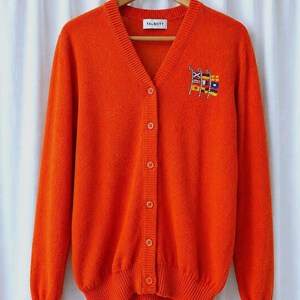 May include: Bright orange cardigan sweater with a V-neck and button-up front. Features a small embroidered design of colorful flags on the left chest. The brand name "TALBOTT" is visible on a small tag at the back of the neck.