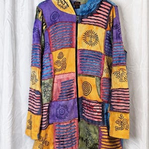 Può includere: Una felpa con cappuccio e zip a maniche lunghe caratterizzata da un design patchwork nei toni del viola, giallo, blu e verde. Il capo presenta vari simboli serigrafati in nero, tra cui soli, spirali e segni Om, oltre a strisce di tessuto testurizzate.