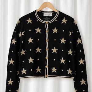 Puede incluir: Un cárdigan negro de manga larga con un patrón repetitivo de estrellas doradas brillantes y pequeños puntos. La prenda tiene un cuello redondo, un cierre frontal con botones dorados y bordes con rayas en color dorado y negro.