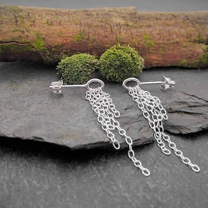 May include: Silver chain earrings with multiple strands hanging from a small hoop. The earrings are displayed on a dark gray, textured surface, with a piece of wood and green moss in the background. The earrings have a simple, elegant design.