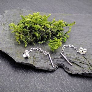 May include: Two silver earring backs with a chain connecting them. The earring backs are on a dark gray slate surface with green moss.