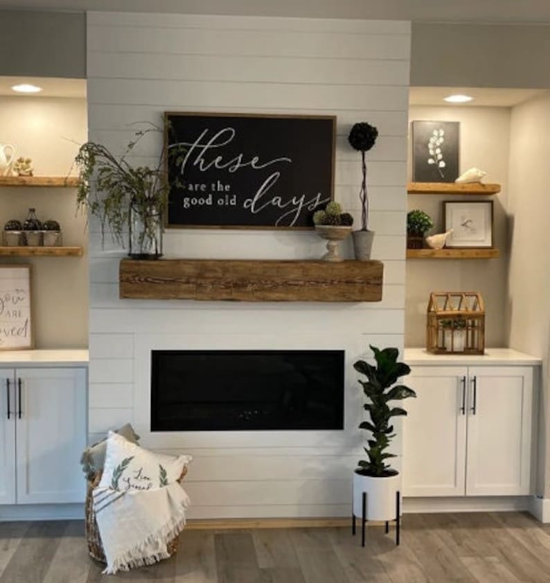 May include: A white fireplace mantel with a black fireplace insert and a wooden beam above. The mantel is decorated with a black framed sign that reads "These are the good old days" and a potted plant. There are also shelves on either side of the fireplace, with decorative items on them.